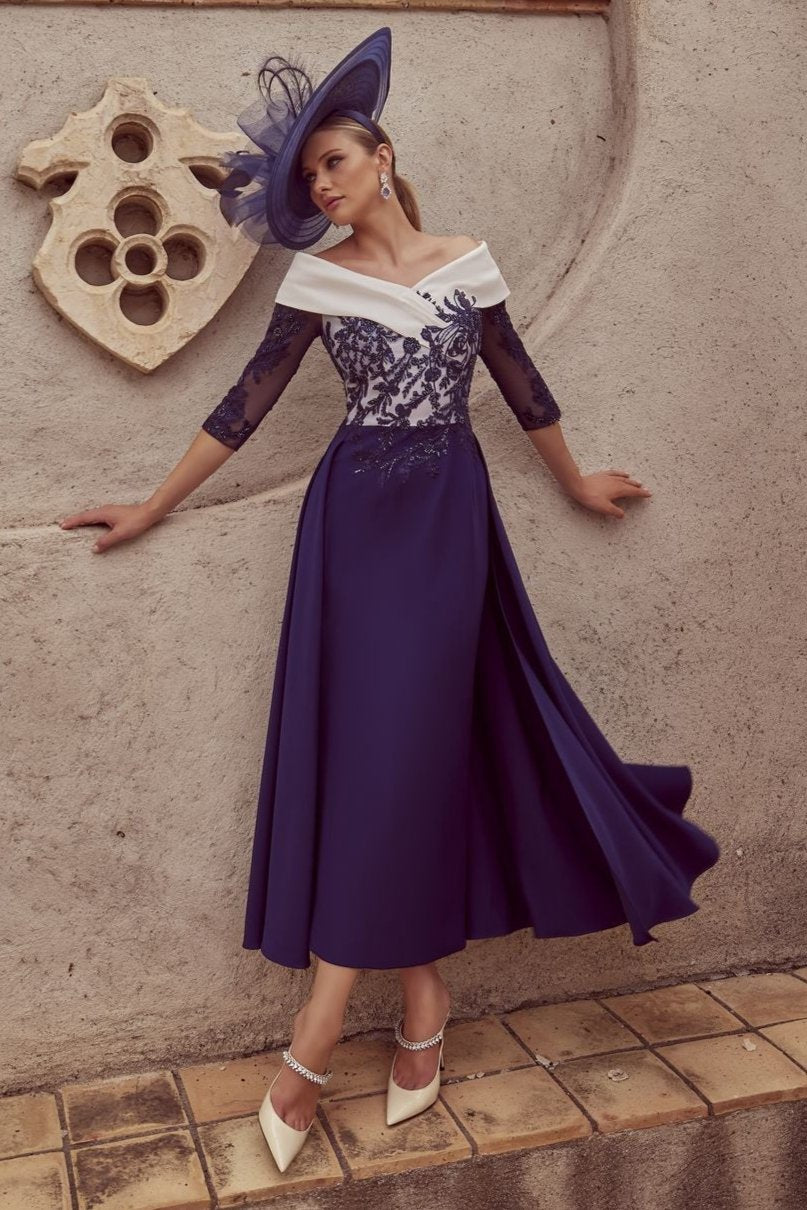 The image showcases a woman posing against a textured gray wall that has a carved decorative element. She is dressed in a full-length navy blue gown with a fitted bodice and lace sleeves, complemented by a wide-brimmed navy hat featuring a feather detail. Her right arm rests casually against the wall, while her left arm extends along her side. Below her, a tiled ledge adds depth to the composition. The palm leaves above create a natural frame, enhancing the overall aesthetic of the scene. The lighting is so