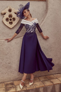 The image showcases a woman posing against a textured gray wall that has a carved decorative element. She is dressed in a full-length navy blue gown with a fitted bodice and lace sleeves, complemented by a wide-brimmed navy hat featuring a feather detail. Her right arm rests casually against the wall, while her left arm extends along her side. Below her, a tiled ledge adds depth to the composition. The palm leaves above create a natural frame, enhancing the overall aesthetic of the scene. The lighting is so