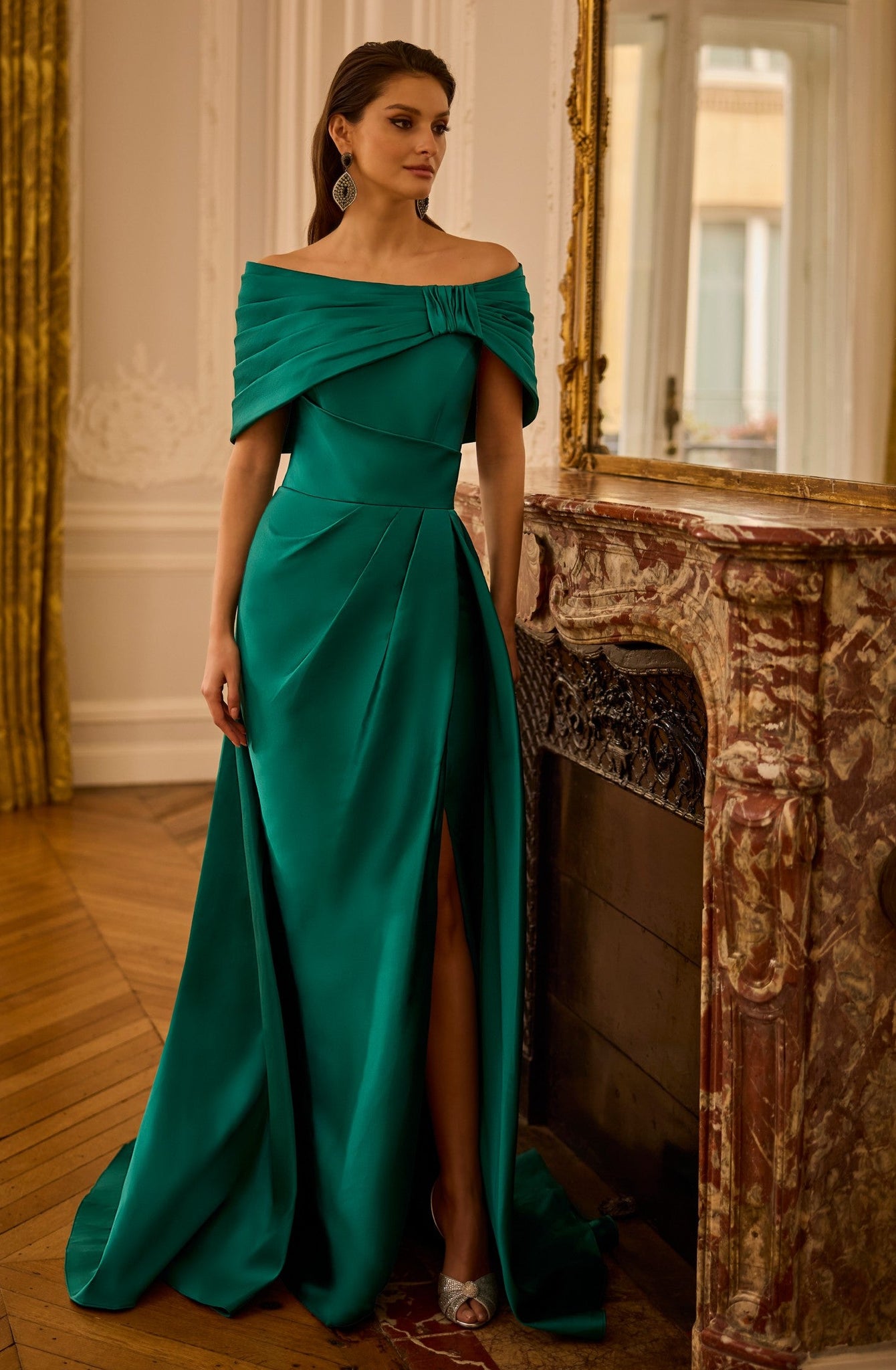Woman in a green off-shoulder dress standing in an elegant room with wooden floor and decorative elements.