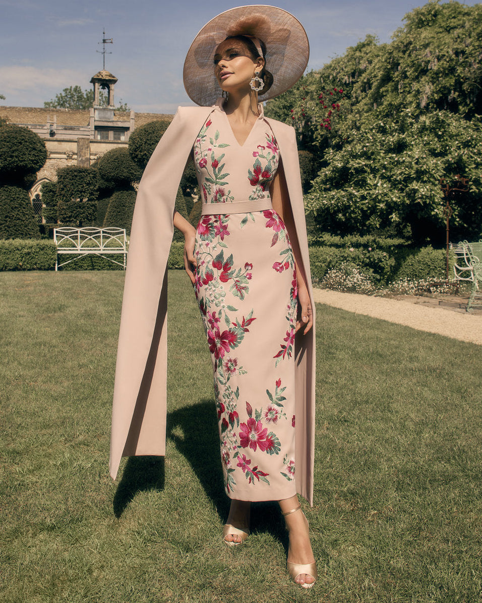 Woman in a floral dress and pink coat standing in a garden with a castle in the background
