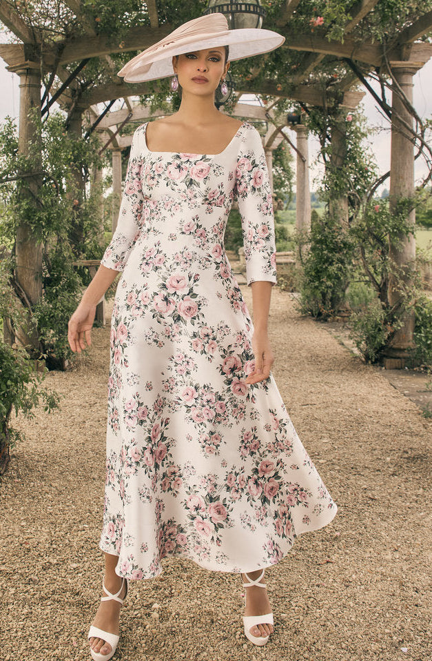 Woman in a floral dress and hat standing under a wooden arbor with greenery.