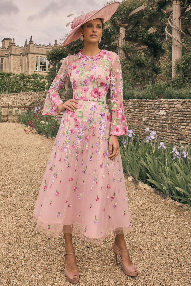 Woman in a floral dress and pink hat standing in a garden with a castle in the background