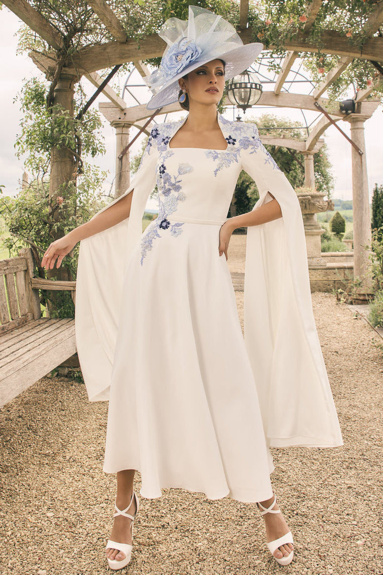 Woman in a white dress with floral accents standing under a wooden arbor in a garden.