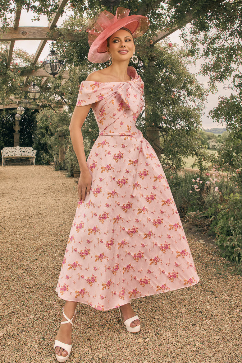 Woman in a pink floral dress and hat standing in a garden setting