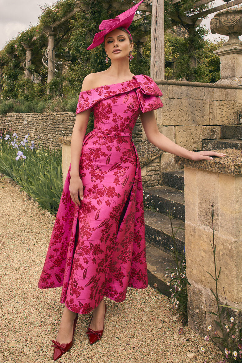 Woman in a pink floral dress and hat standing in an outdoor garden setting.