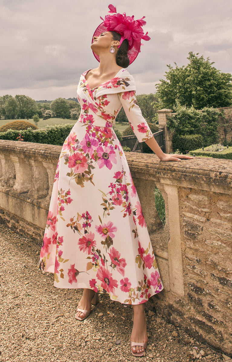 Woman in a floral dress and pink hat standing on a stone wall with a garden and trees in the background.