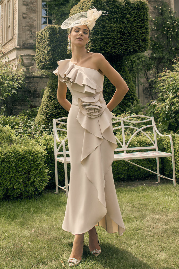 Woman in a beige dress and hat standing in a garden with a castle-like building in the background