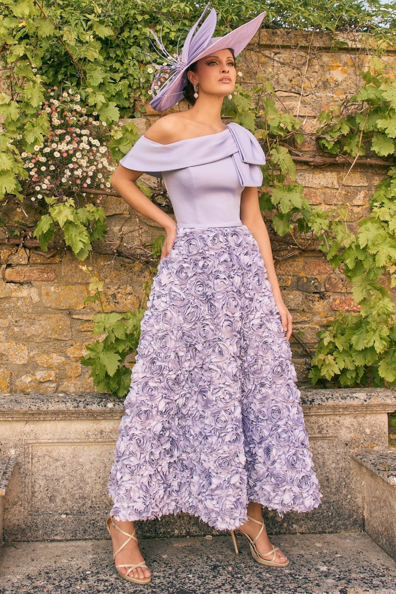 Woman in a lavender outfit with a matching hat standing in front of a stone wall with greenery.