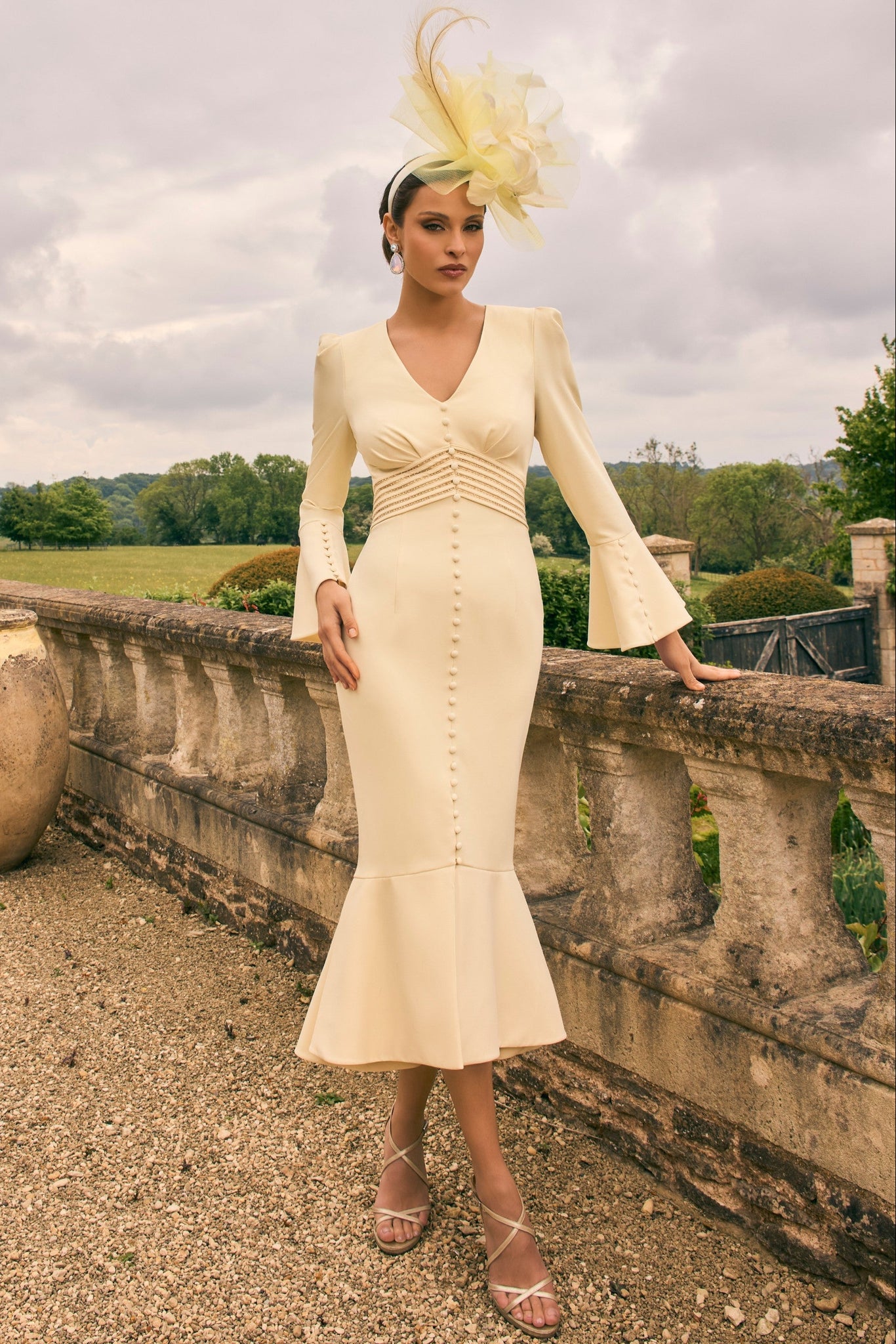 Woman in a cream dress with a large decorative hat standing on a stone balcony with a scenic background.