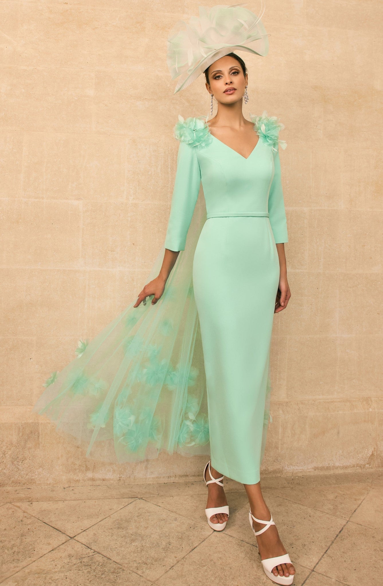 Woman in a mint green dress with ruffled sleeves and a matching headpiece against a beige wall.