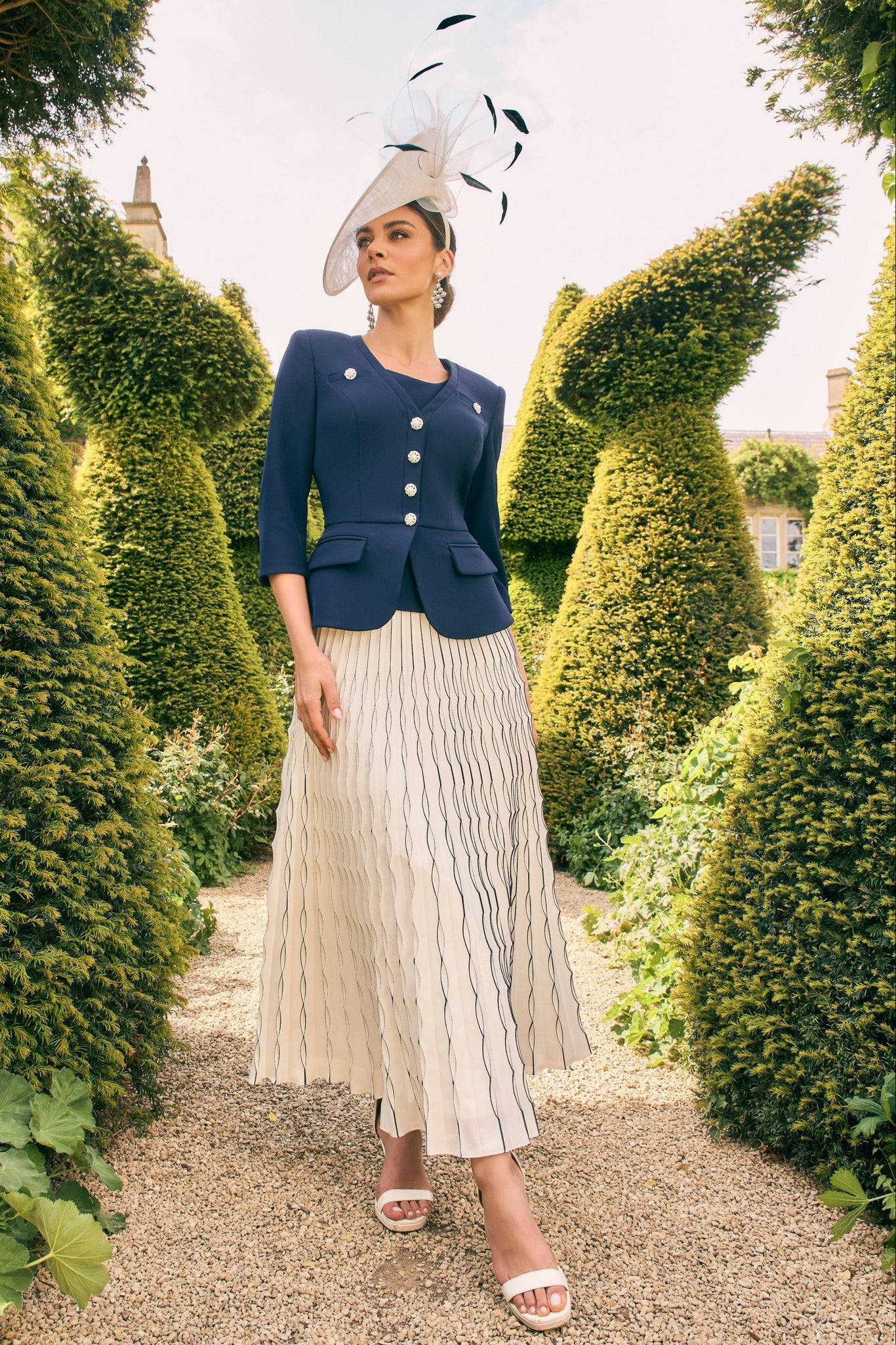 Woman in formal attire with a navy blazer and white skirt standing in a garden with topiary.
