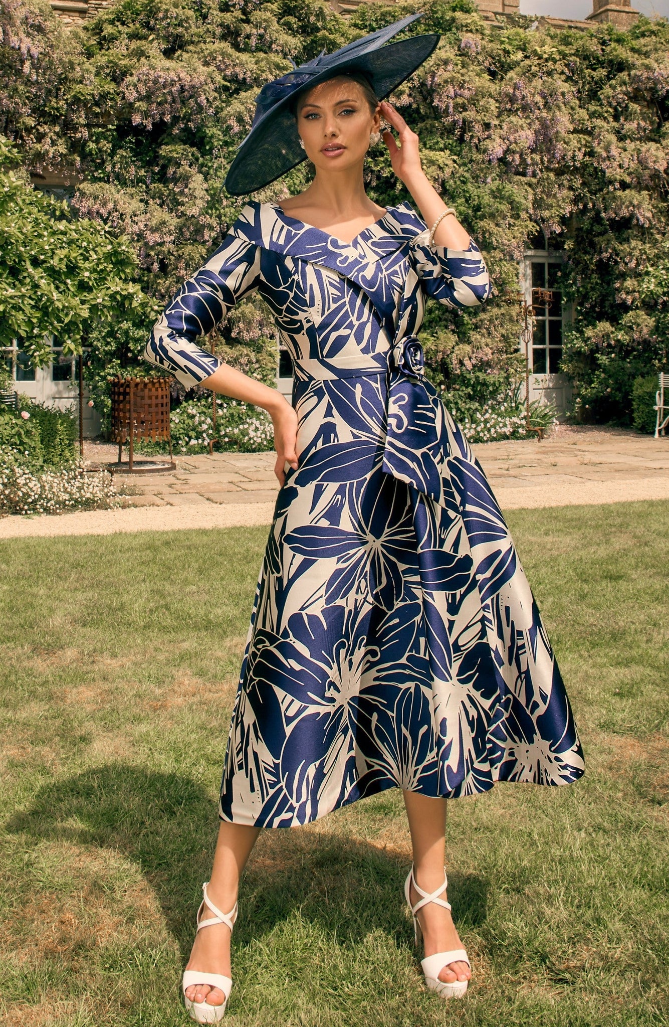 Woman in a blue floral dress and large hat standing in a garden.