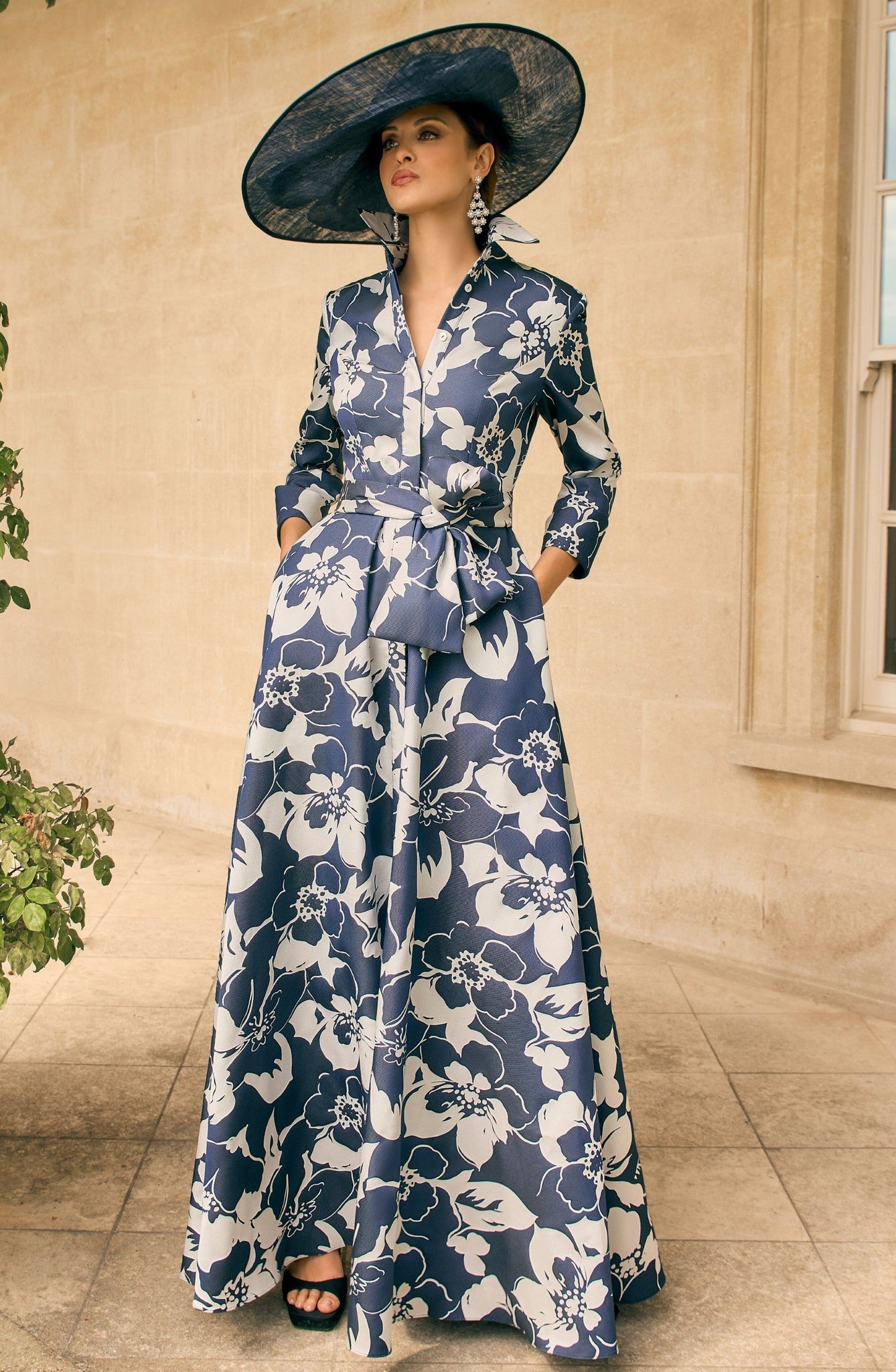 Woman in a blue floral dress standing in a sunlit room with a window and plant.