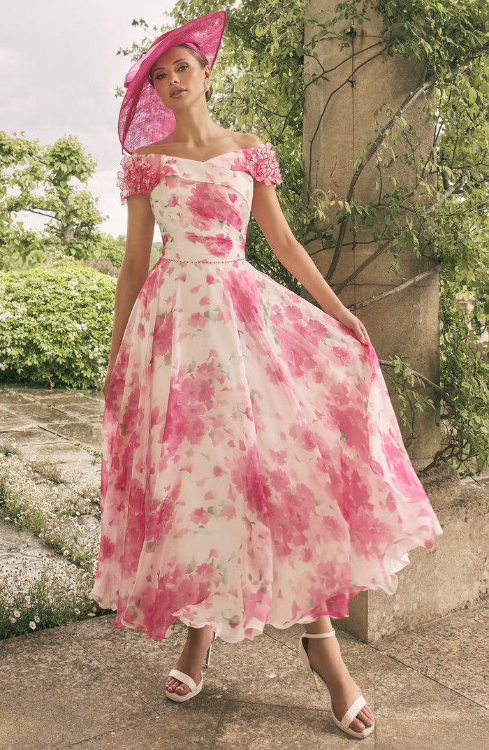 Woman in a pink floral dress and hat standing in a garden.