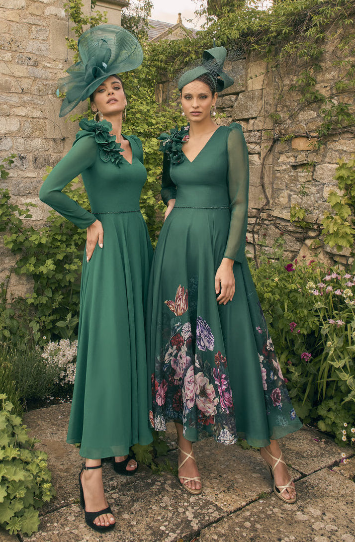 Two women in green dresses standing in a garden with stone walls and plants.