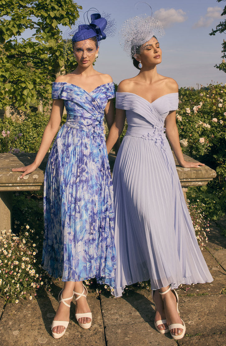 Two women in floral and plain dresses standing outdoors with greenery and a clear sky.