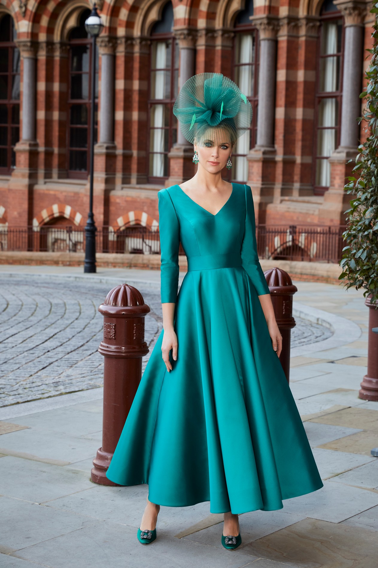 Blonde lady standing outside St Pancras Hotel London wearing A-line occasion dress with full skirt and v-neckline and matching court heel shoes and fascinator headwear accessory