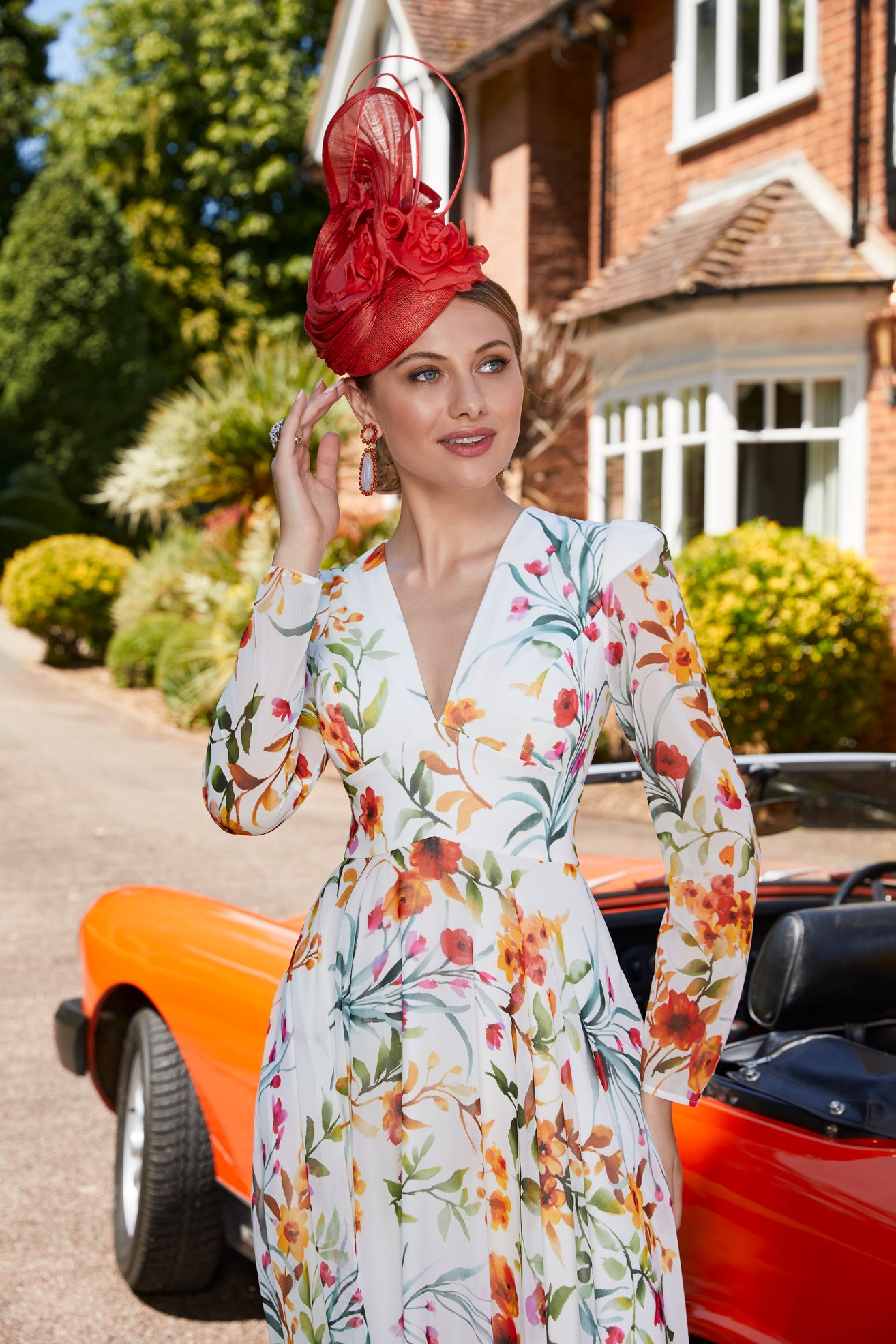 Woman standing next to vintage orange car in front of house wearing bold floral print long sleeve special occasion dress and fascinator head accessory