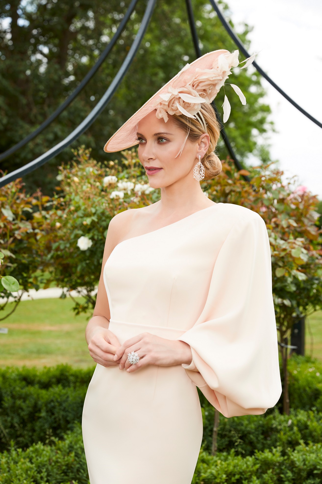 Close up image of a blonde woman in one shoulder blouson sleeve special occasion dress in matching fascinator hat standing in a garden in front of plants