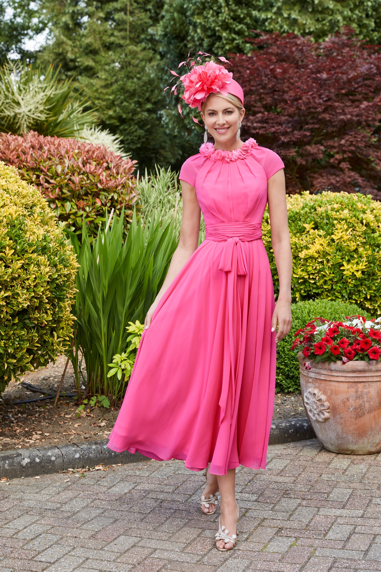 Lady in garden wearing vibrant pink a-line dress with 3d floral neckline, gathered waist detail and cap sleeves with matching occasion headwear piece